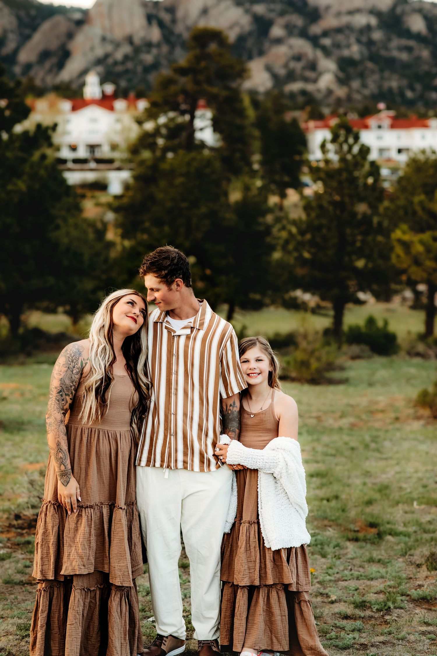 A family in coordinated brown and white outfits poses together against a mountain backdrop with white buildings.