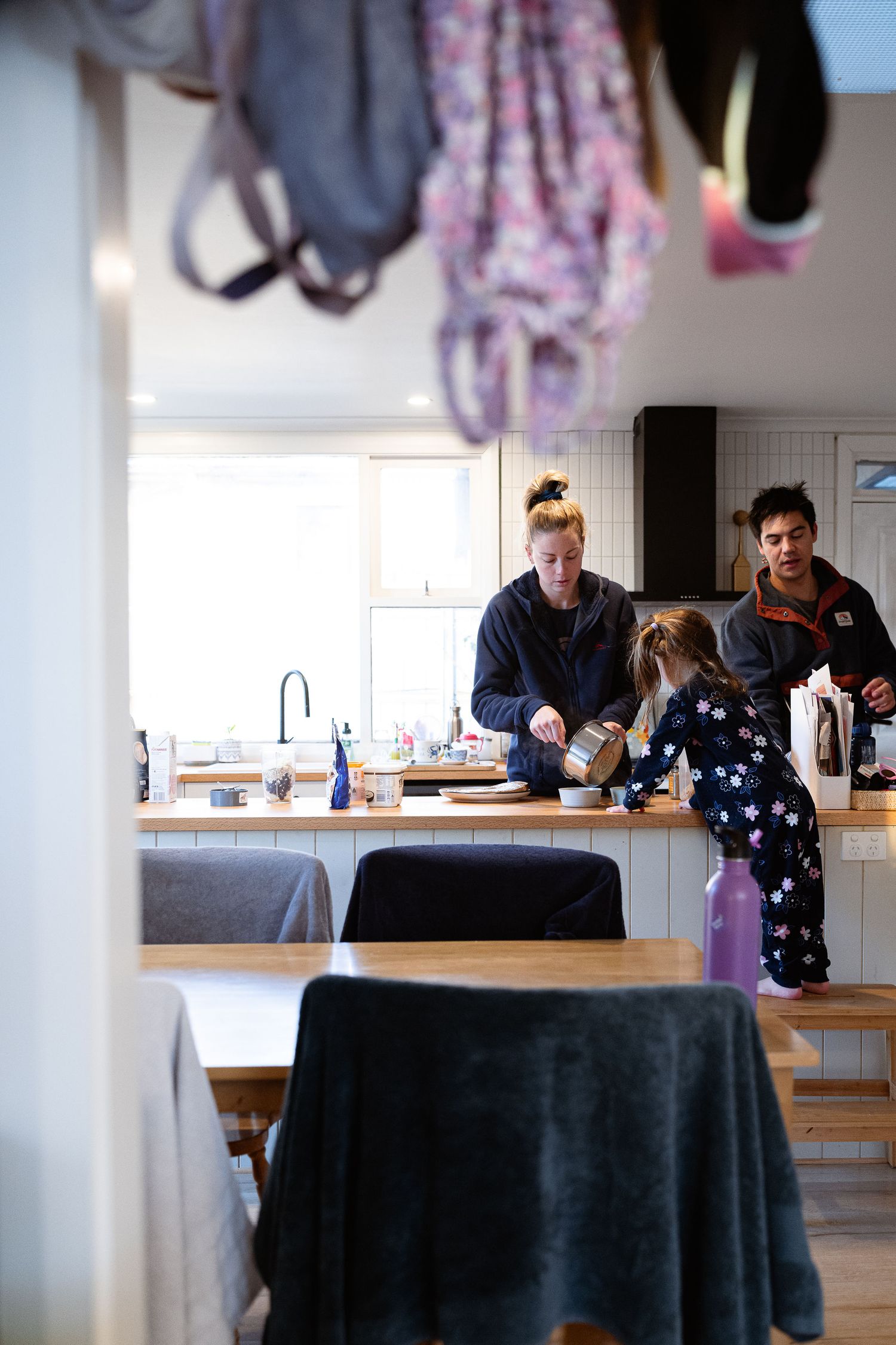 People gather in a kitchen with dining area, featuring dark tablecloths and hanging clothing in the background.
