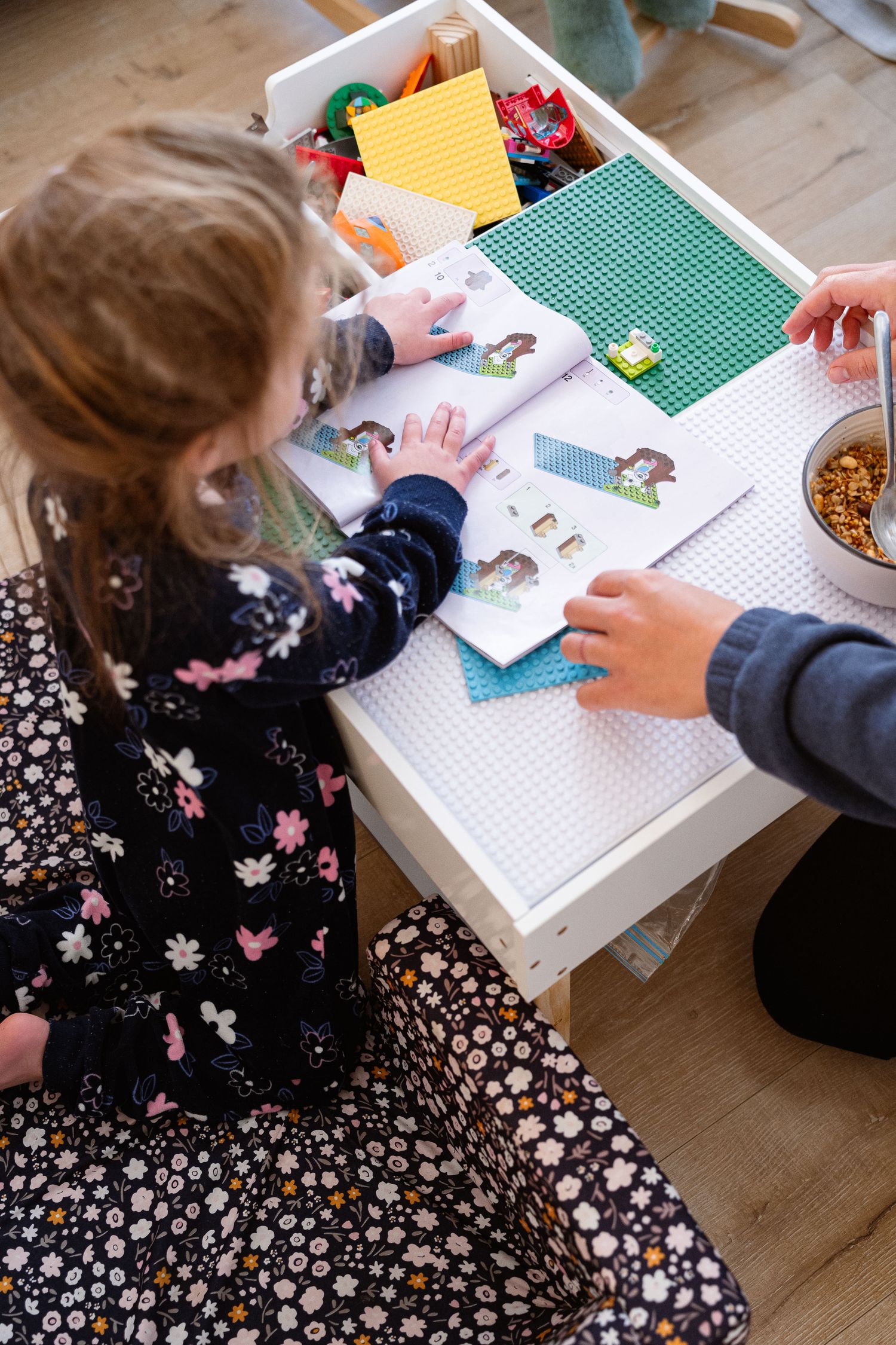 Two people playing with stickers and toys at a white table with a green baseplate and craft supplies.