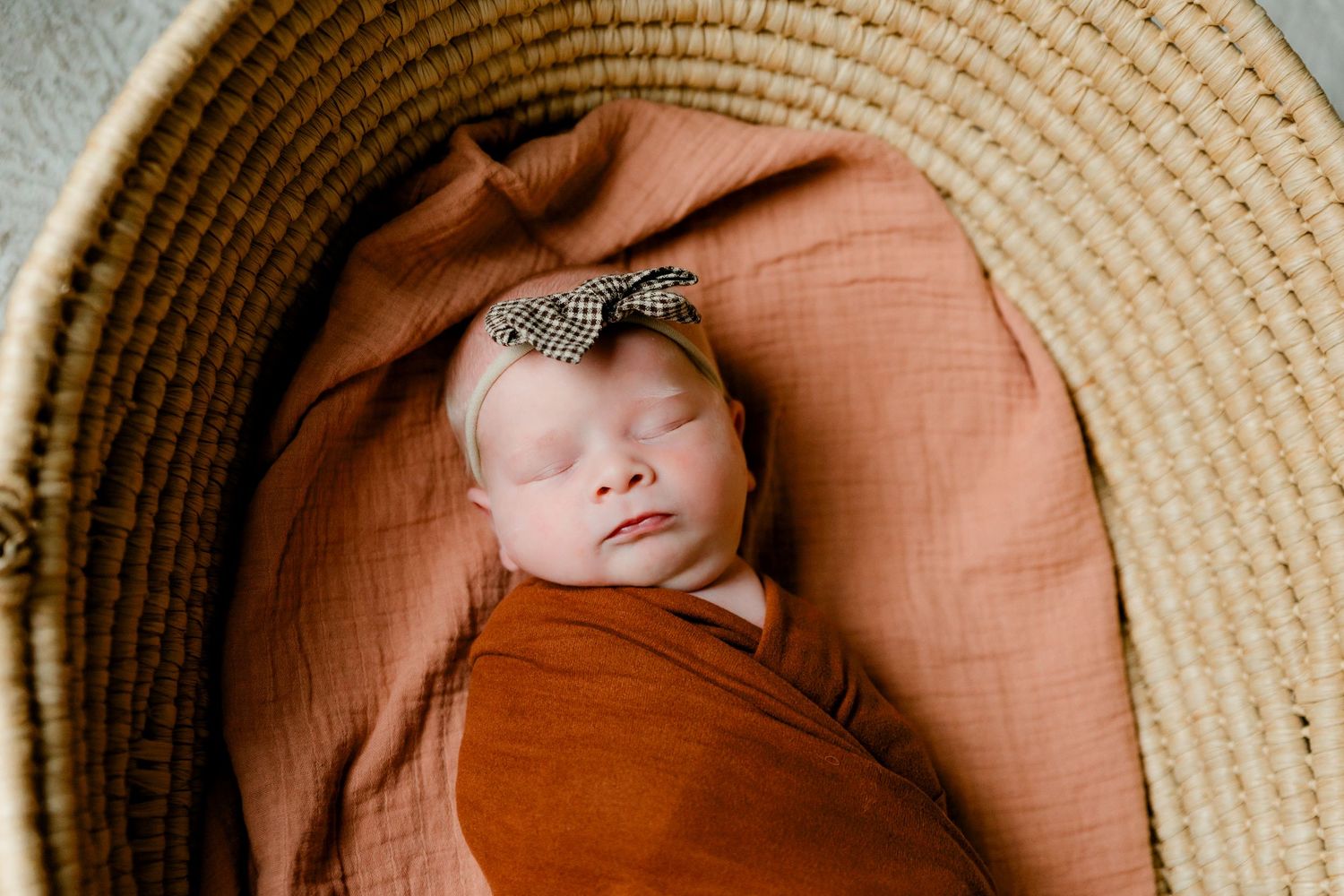 Sleeping newborn baby girl wrapped in rust orange swaddle with bow headband in woven basket.