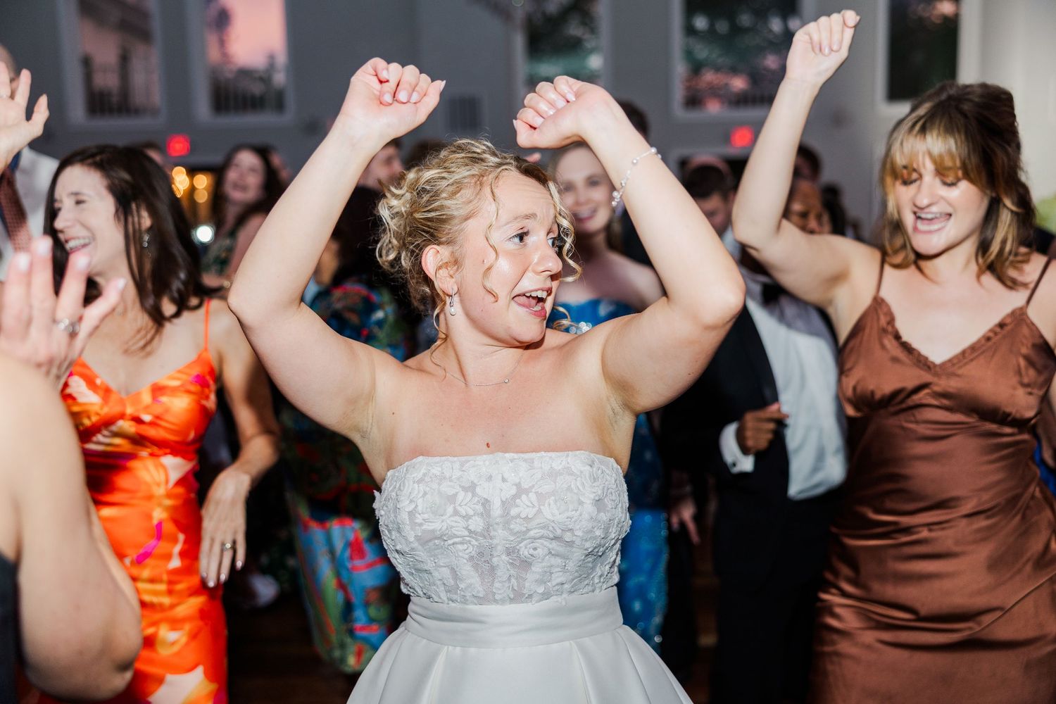 Excited wedding guests dance with arms raised on a wooden dance floor during reception celebrations.