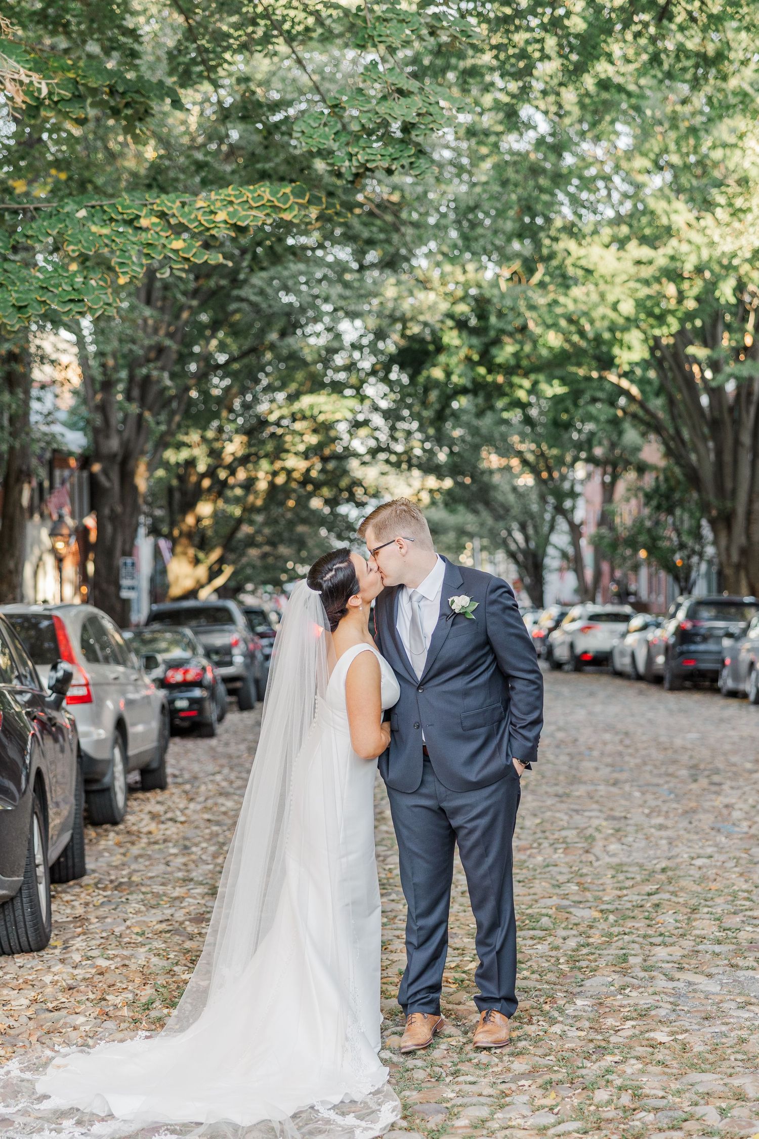 Couple in wedding attire shares romantic moments on a tree-lined street covered in fallen autumn leaves.