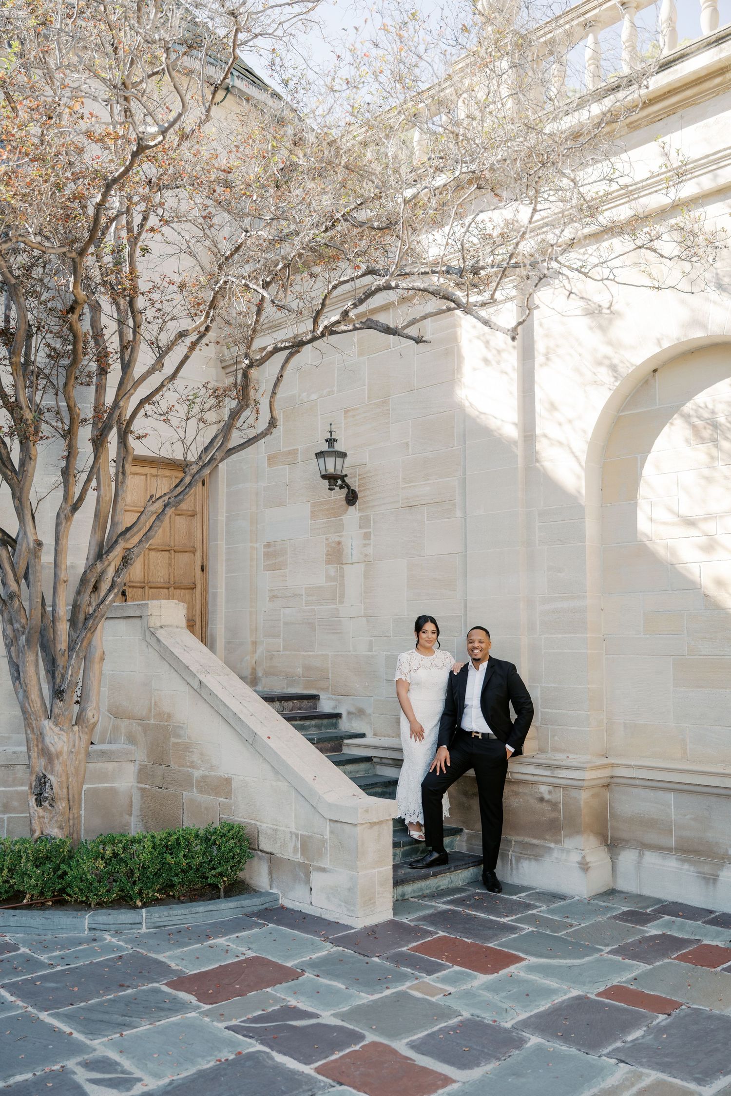 A couple stands on elegant stone steps beside a blooming tree at an ornate architectural entrance.