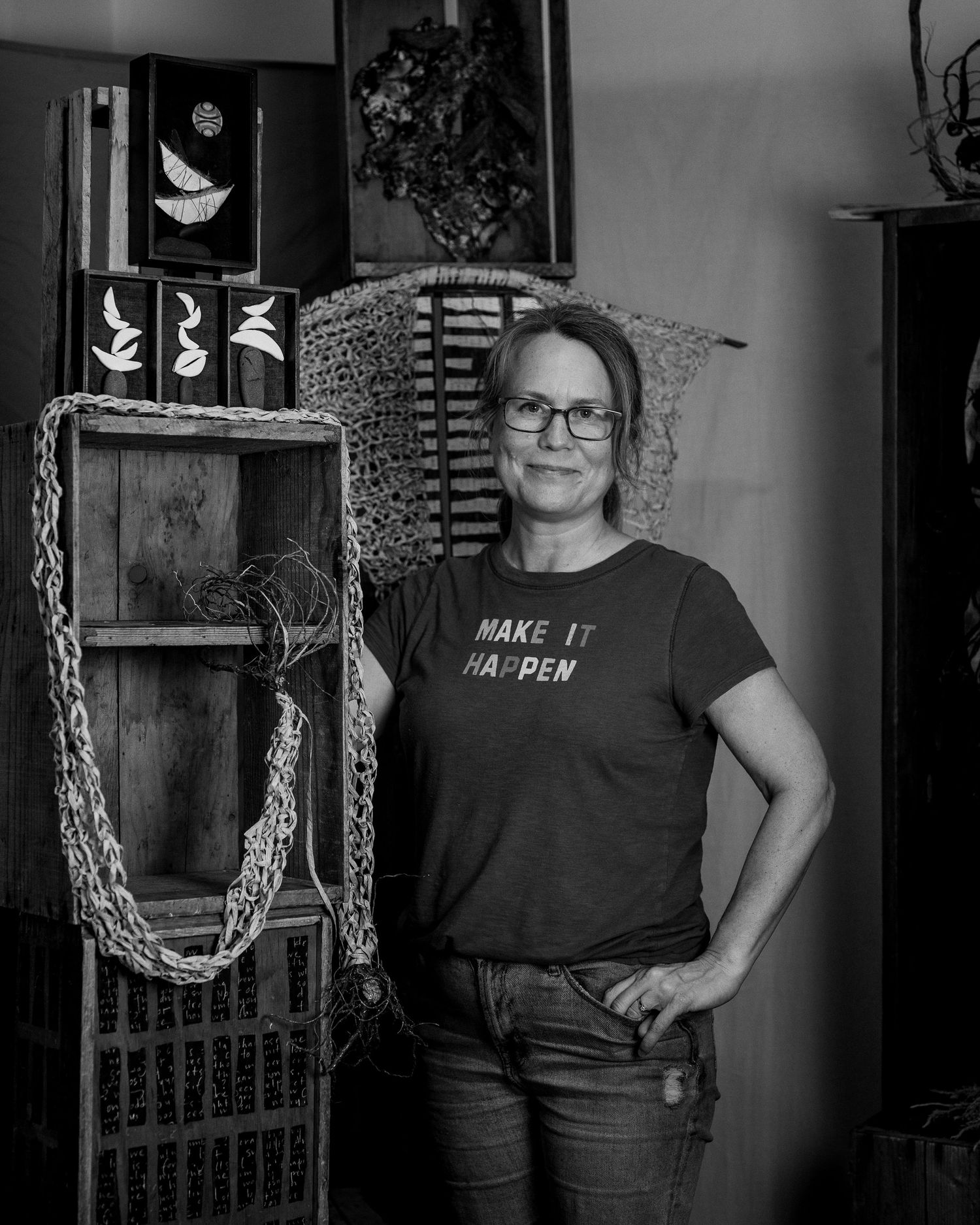 A black and white photograph showing someone in a "Make it happen" t-shirt standing next to rustic wooden shelving with decor.
