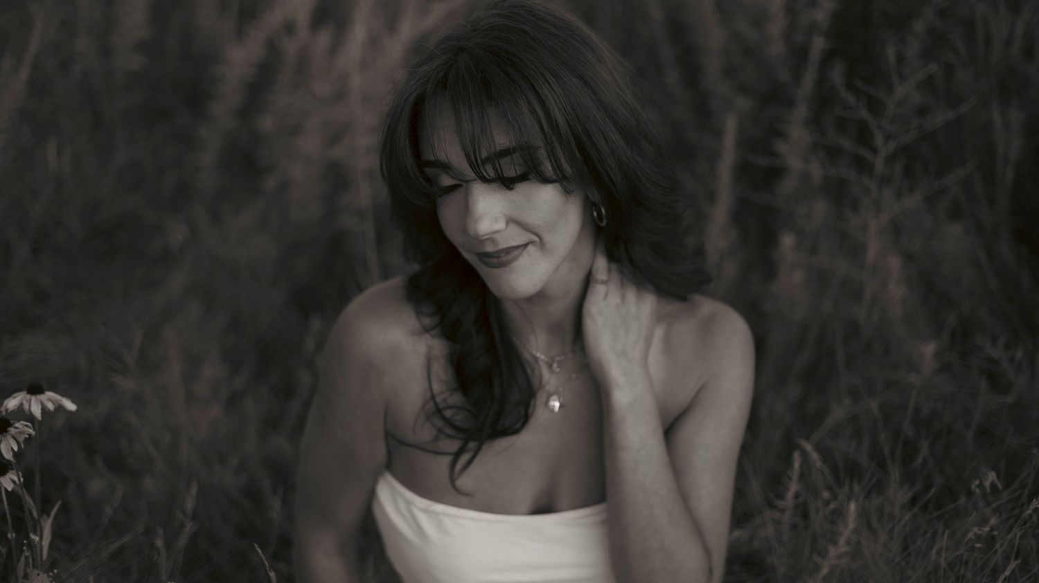 Black and white portrait of a woman in a strapless dress sitting outdoors among tall grass.