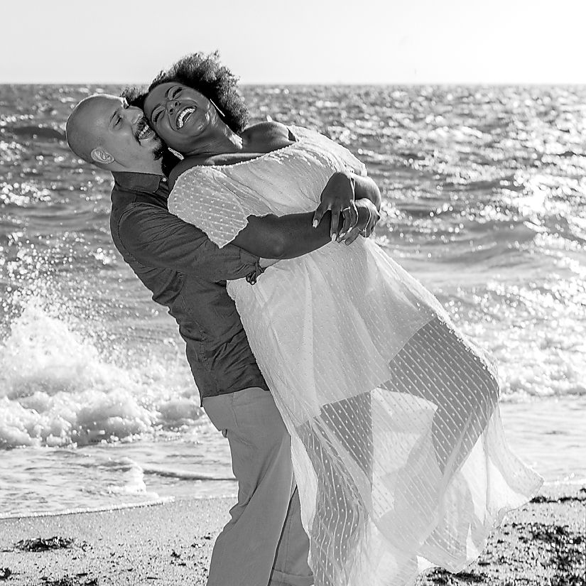 A joyful couple embraces on the beach in black and white as waves crash behind them.