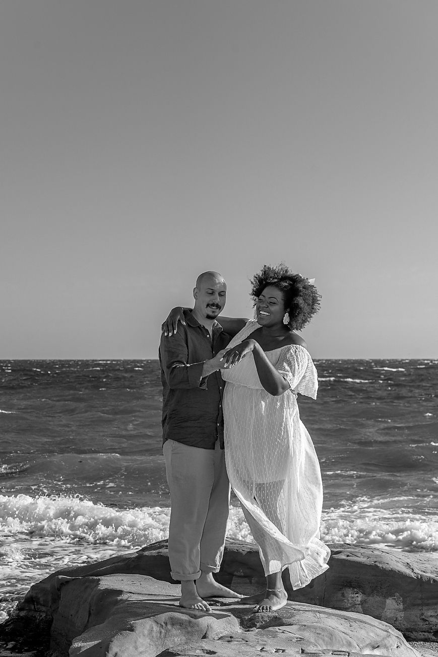 A couple stands together on rocky coastal outcrop with crashing waves and ocean in the background.