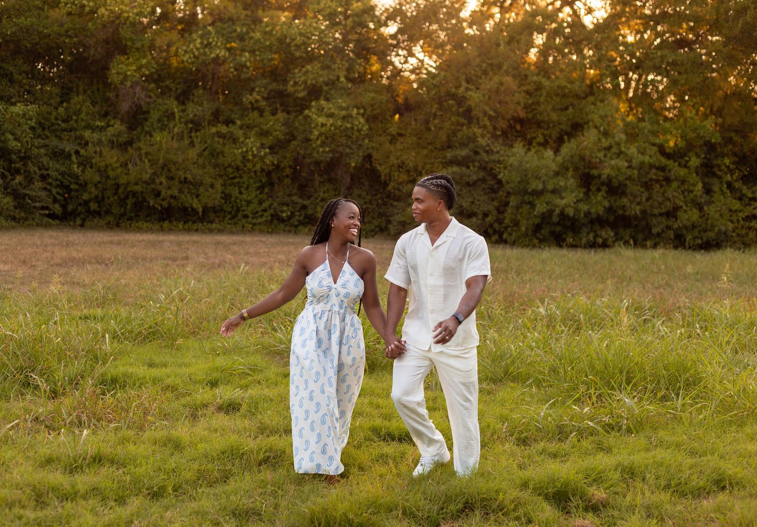 A couple in white outfits walks together in a grassy field during golden hour sunset.