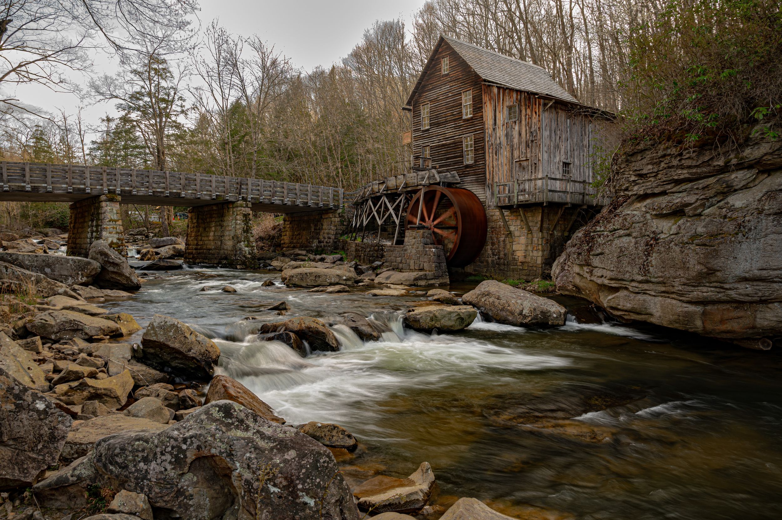 Historic Babcock grist mill and wooden bridge spanning a rushing river surrounded by rocky terrain.
