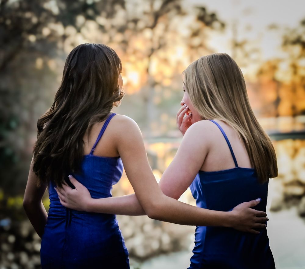 Two people in navy blue dresses embrace while standing outdoors during golden hour sunset.