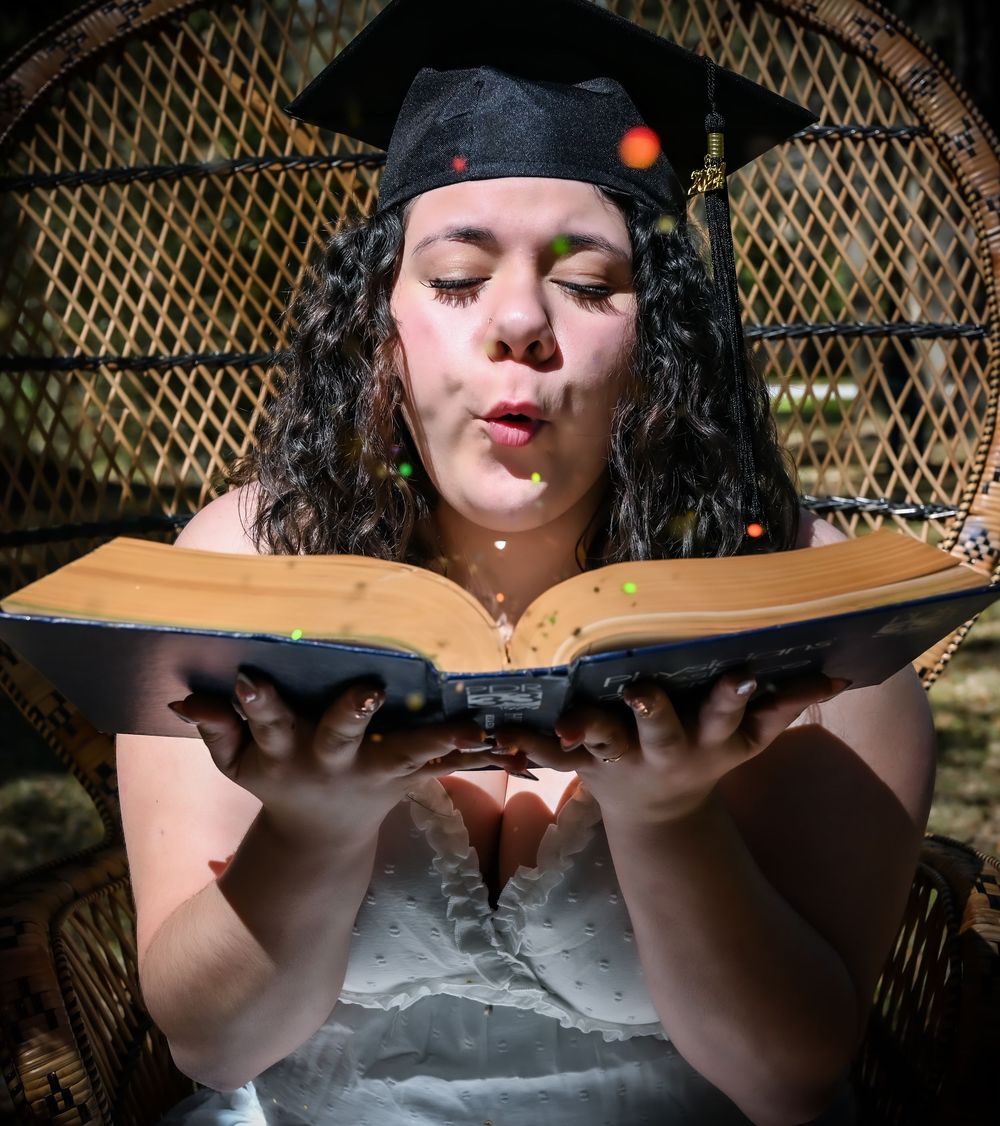 A person in a black headpiece blows glitter from an open book while seated in a wicker peacock chair.