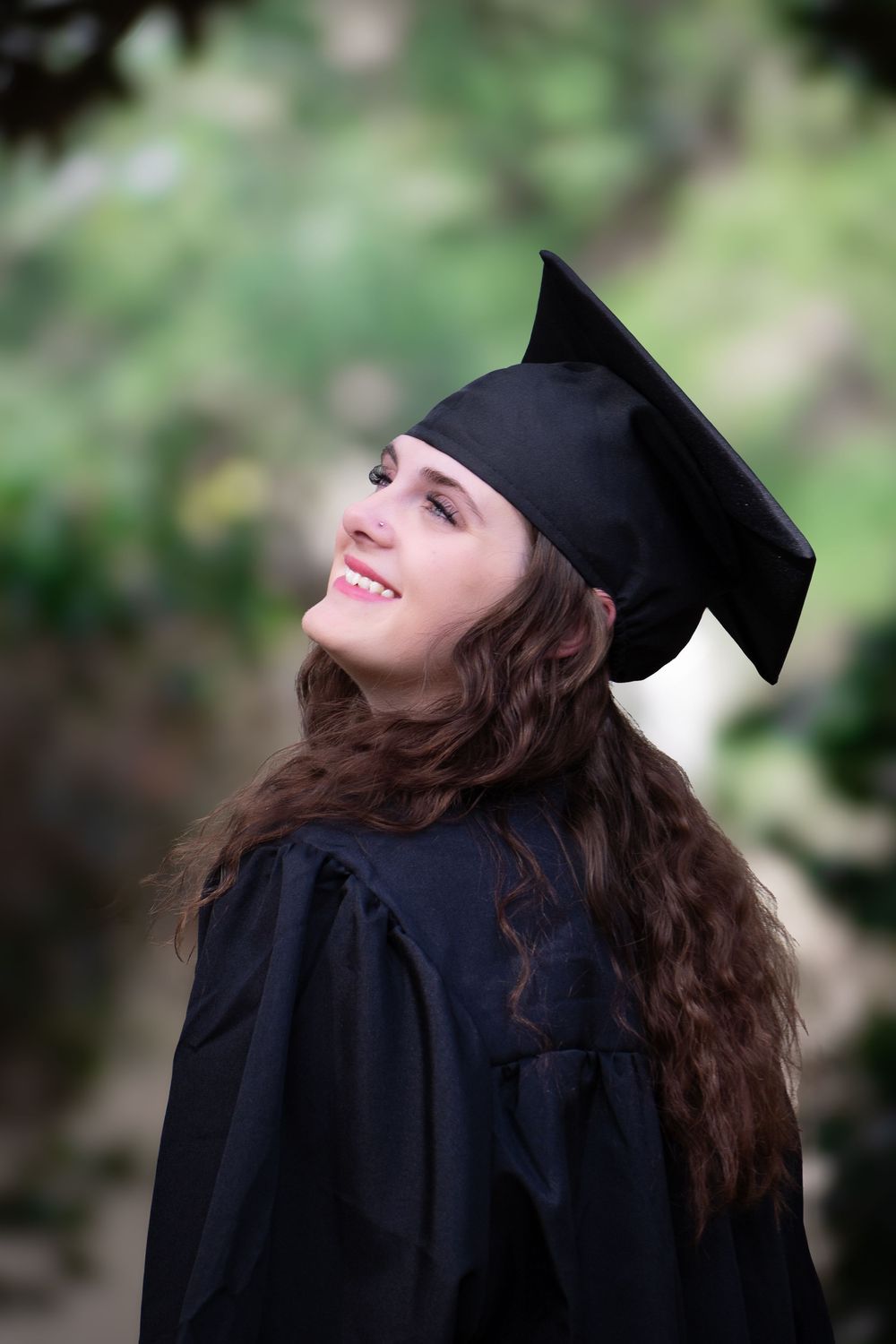 Graduate in black cap and gown smiles joyfully while looking up during outdoor graduation photoshoot.