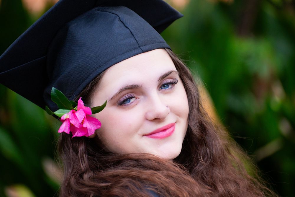 Graduate in black cap with pink flower smiles warmly during outdoor graduation photo session.