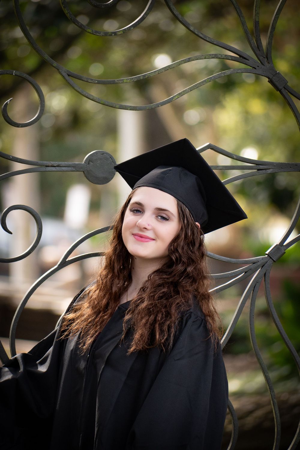A graduate in cap and gown poses outdoors next to decorative iron scrollwork with a soft bokeh background.