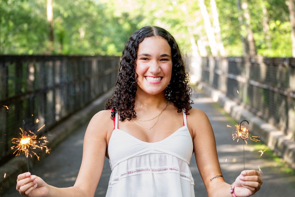 A person in a white dress holds sparklers in a tree-lined pathway during a summer celebration.