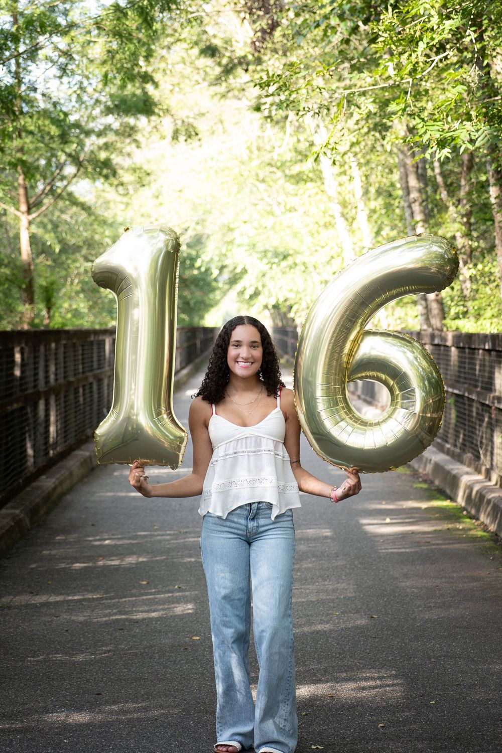 Person holding large metallic number balloons on a tree-lined pathway.