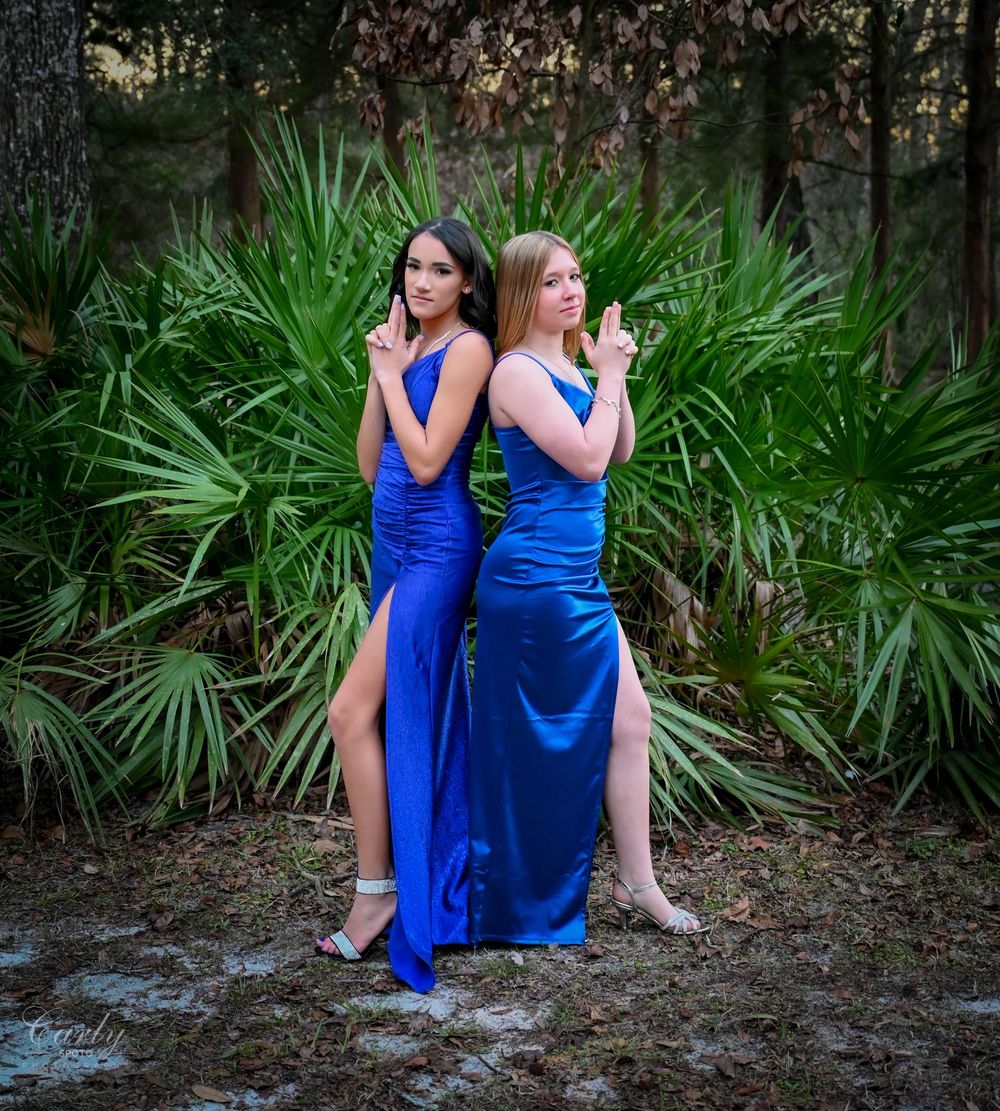 Two people in royal blue dresses sit together on natural ground surrounded by lush green palm fronds.