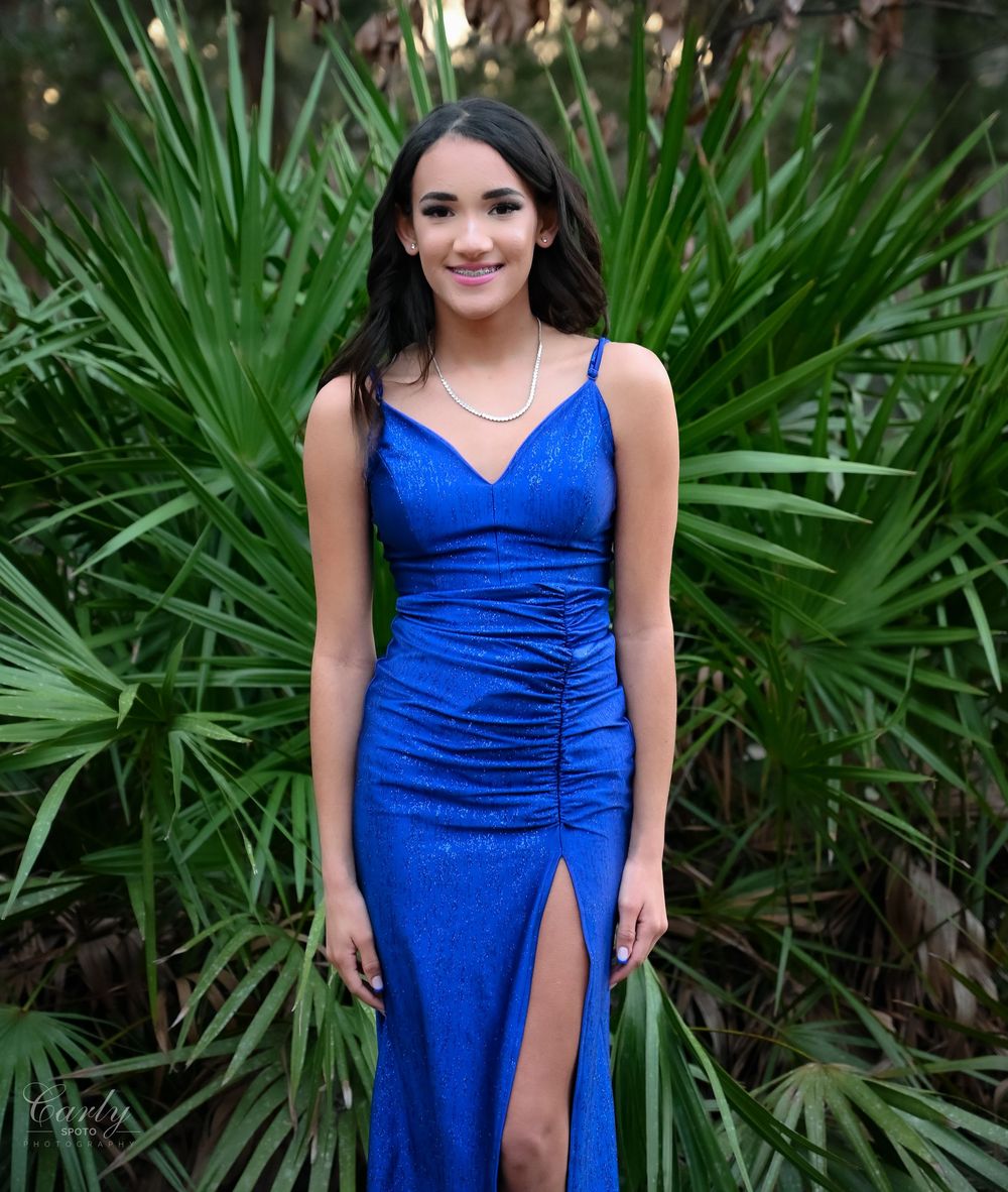 A person in a royal blue evening gown poses elegantly against a backdrop of lush tropical palm fronds.