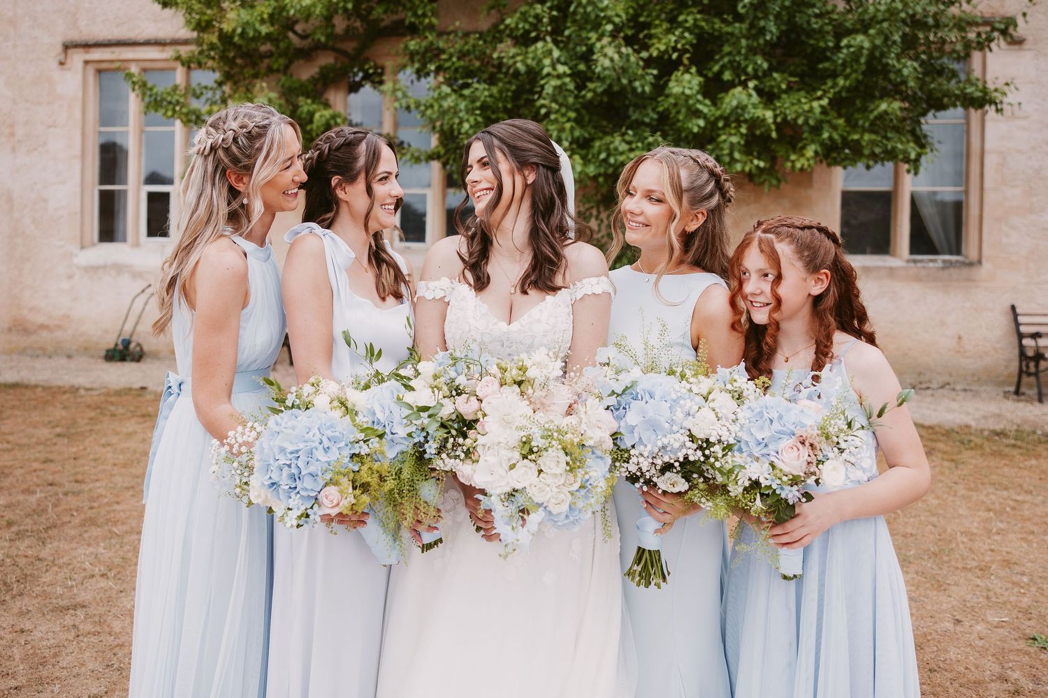 Row of bridesmaids in light blue dresses holding white floral bouquets pose together at outdoor wedding venue.