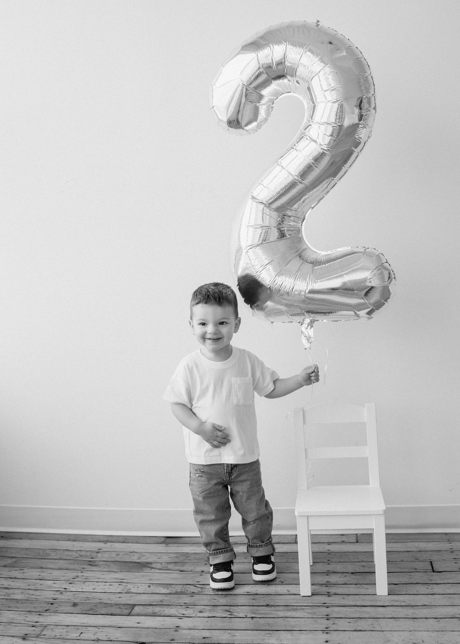 A toddler in a white shirt and jeans stands next to a white chair with a silver number 2 balloon in a minimalist setting.