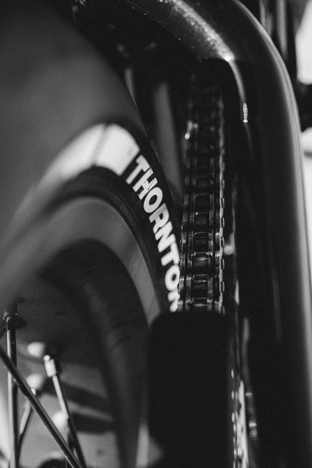 Close-up black and white detail shot of a bicycle chain and Thornton branded wheel in dramatic lighting.