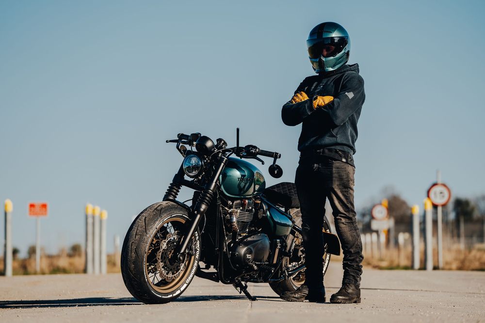 A motorcyclist in black riding gear sits on a parked motorcycle against a desert landscape with poles in the background.