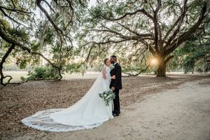Romantic wedding couple embraces under massive oak trees draped with Spanish moss at sunset.