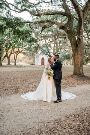 Newlyweds embrace on path beneath Spanish moss-covered oaks with white chapel in background.