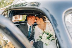 A romantic moment captured through the window of a vintage car with white flowers and formal attire.