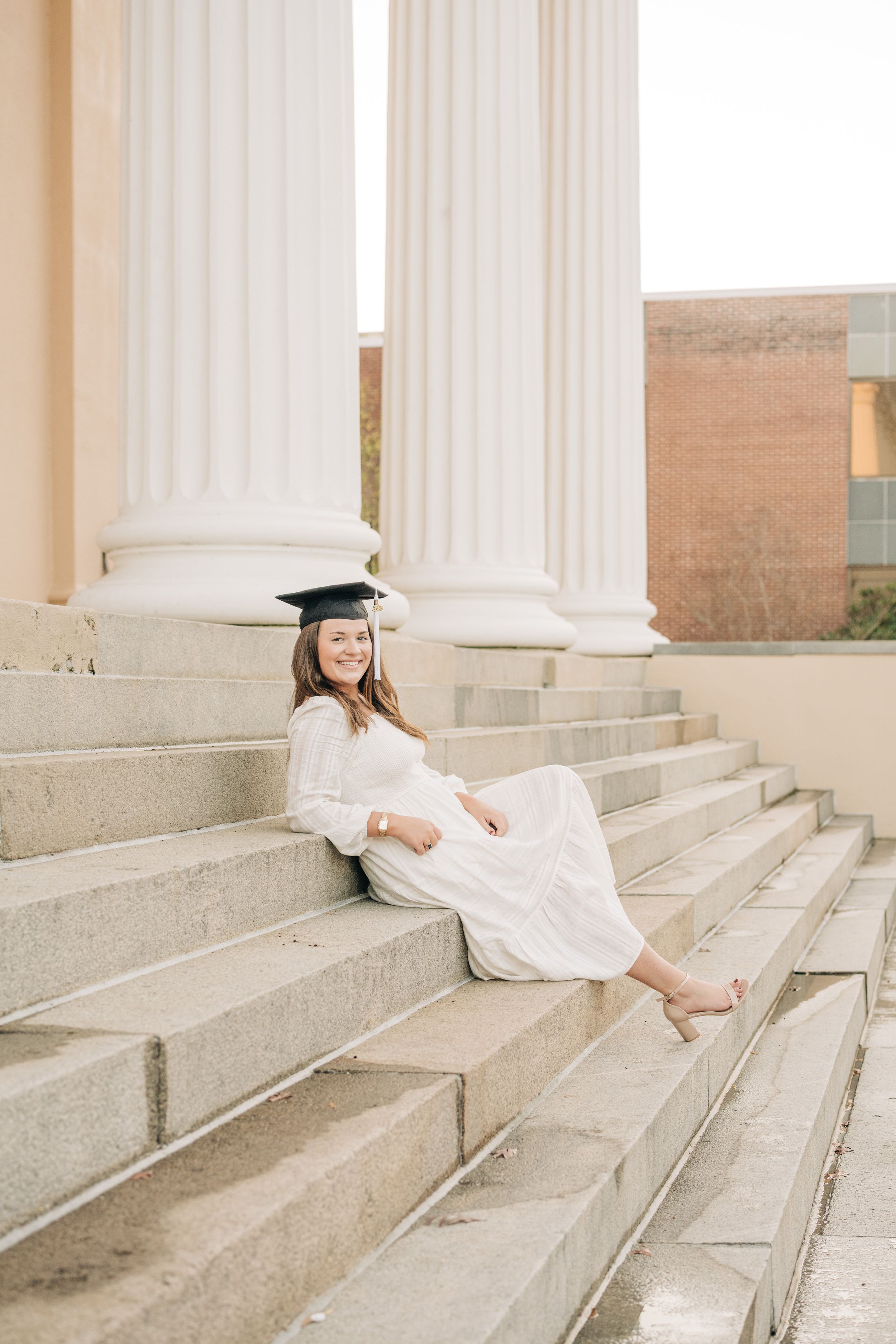 Graduate in white dress and cap sits on stone steps near classical columns in soft natural lighting.