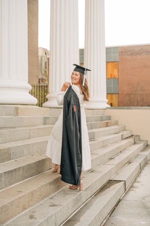 Graduate in cap and gown stands on stone steps with white columns in background during graduation photoshoot.