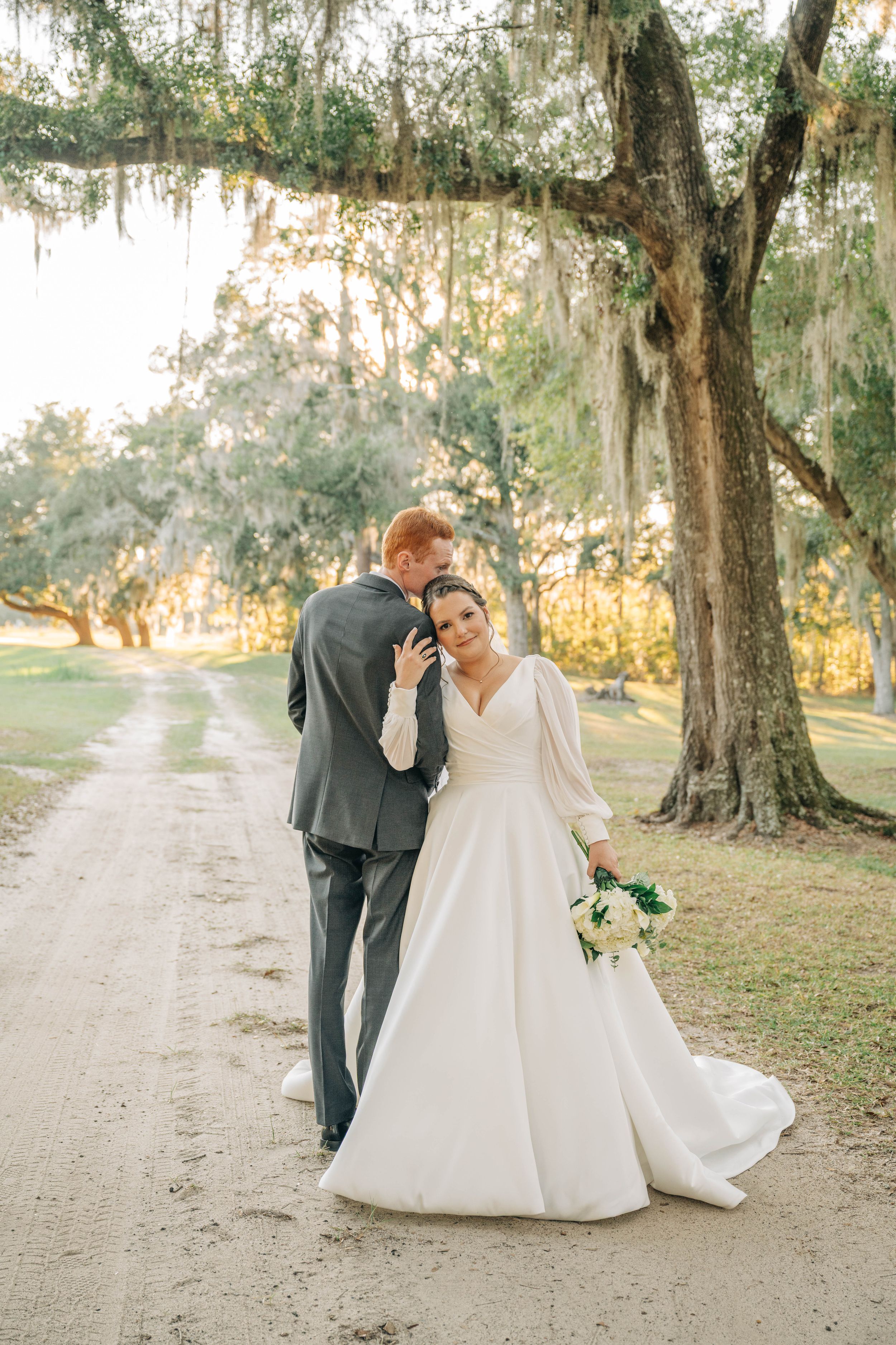 A romantic wedding couple walks along a dirt path under Spanish moss-draped oak trees at sunset.