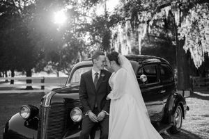 Vintage black car parked under Spanish moss draped trees at a wedding ceremony in a park setting.