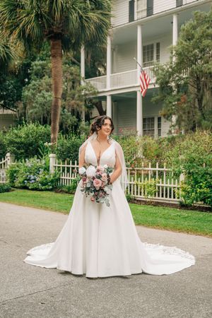 A bride in a white wedding gown poses with a pink and white bouquet on a paved path near a colonial-style building.
