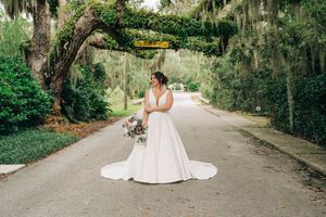 A bride in a flowing white gown poses on a tree-lined pathway in a romantic garden setting.