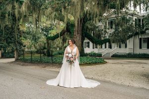 A bride in a flowing white wedding gown holds a bouquet while standing on a path beneath Spanish moss-draped trees.