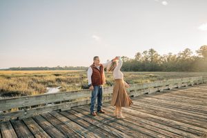 Two people walk along a wooden boardwalk through a golden marsh at sunset creating a romantic silhouette.