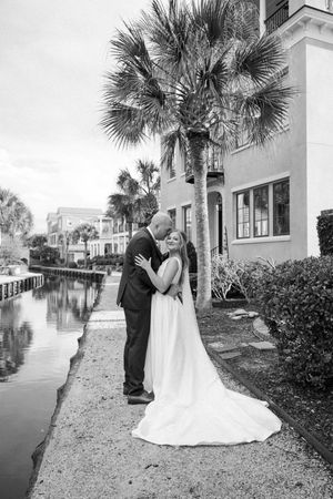 A romantic wedding photoshoot along a palm-lined waterfront path with white buildings in the background.