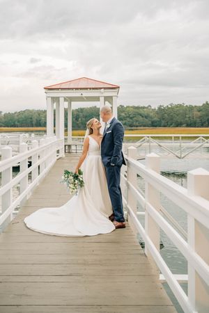 A wedding couple sharing an intimate moment on a white wooden dock overlooking a serene waterway.