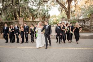 Wedding party in black attire poses playfully behind a couple on a historic brick walkway.