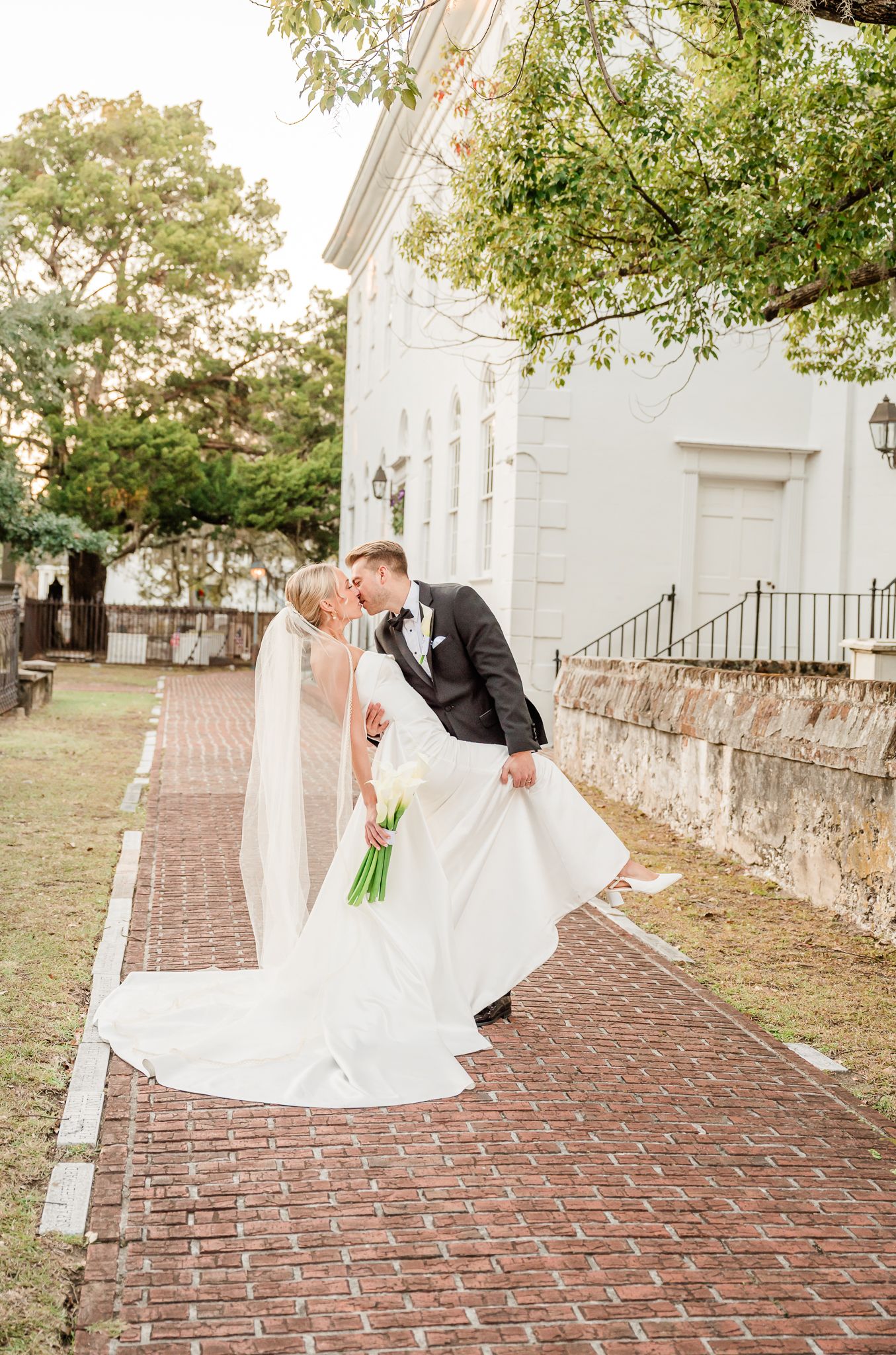 Elegant wedding couple poses on brick pathway beside white historic building.