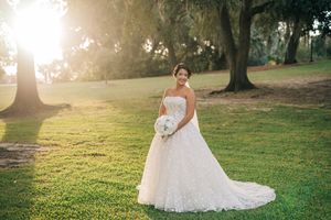 A bride in a white lace wedding dress poses in a sunlit garden with trees and golden light creating a dreamy atmosphere.
