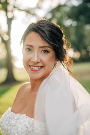 A radiant bride wearing a white strapless lace wedding dress and veil smiles in warm sunlit outdoor setting.
