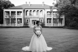 A bride in a white wedding gown stands on a grand lawn in front of a stately mansion with white columns.