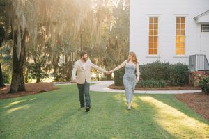 A couple holds hands while walking on a sunlit lawn in front of a white church with Spanish moss in the background.