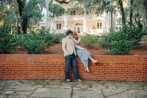 A romantic couple shares a tender moment on a brick wall with a stately Southern mansion and Spanish moss in the background.