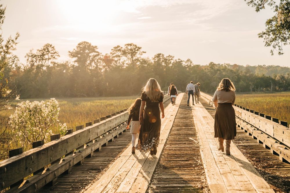 People walking on a wooden boardwalk at sunset with golden light streaming through trees in a natural setting.