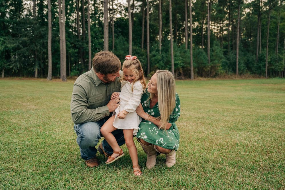 A family enjoys a playful moment together on a grassy lawn with pine trees in the background.