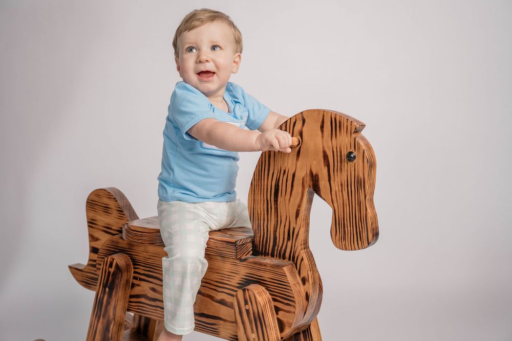 A toddler in light blue shirt and white pants stands on a wooden rocking horse against a gray background.