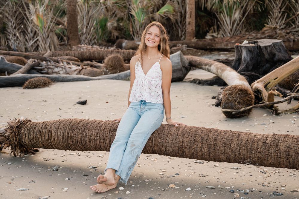 Beachside lifestyle photo shoot featuring someone in casual denim and white top sitting on driftwood logs at sunset.